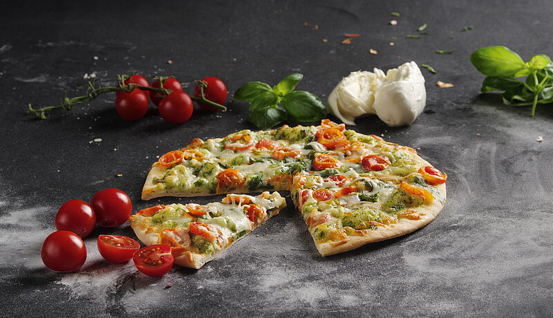 Aufgeschnittene Pizza mit Tomate, Spinat und Käse, ein Stück fehlend. Drumherum Tomaten, Bailikum und Knoblauch drapiert.