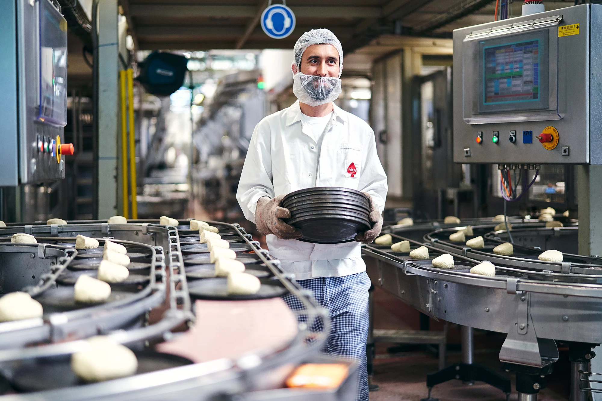 Mitarbeiter läuft am Fließband mit Pizzateigen vorbei und hält gestapelte Bleche in der Hand.