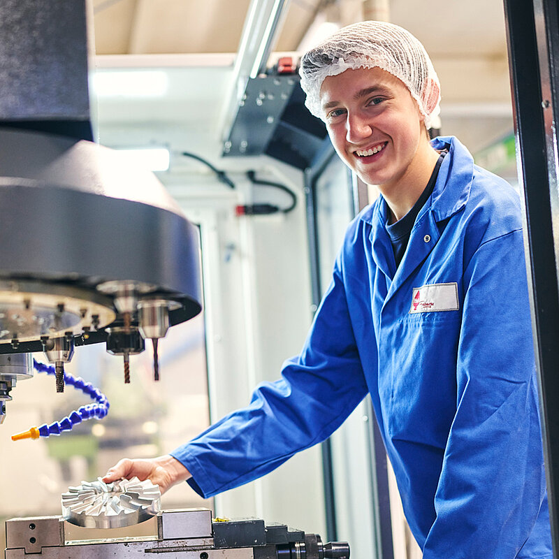 Ein Mitarbeiter in Arbeitskleidung steht lächelnd vor einer Maschine.