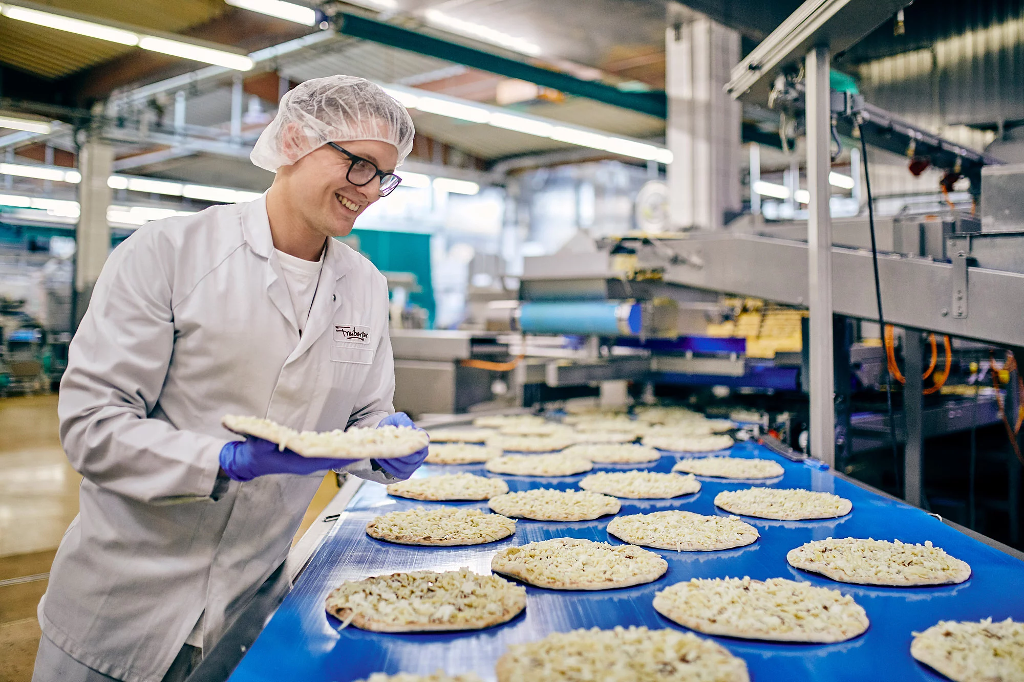 Mitarbeiter in Arbeitskleidung steht vor einem Fließband befüllt mit Pizzen und hält lächelnd eine Pizza in der Hand.