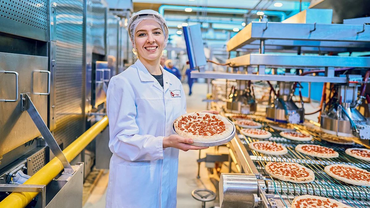Mitarbeiterin in Arbeitskleidung am Produktionsband hält eine Pizza lächelnd in die Kamera. 