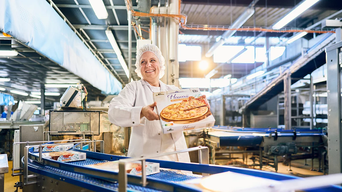 Mitarbeiterin in Arbeitskleidung mit Pizzakarton in der Hand an der Verpackungsmaschine.