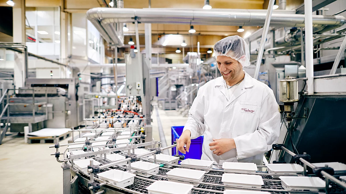 Mitarbeiter in Arbeitskleidung lächelnd am Band von abgepackten Gerichten.