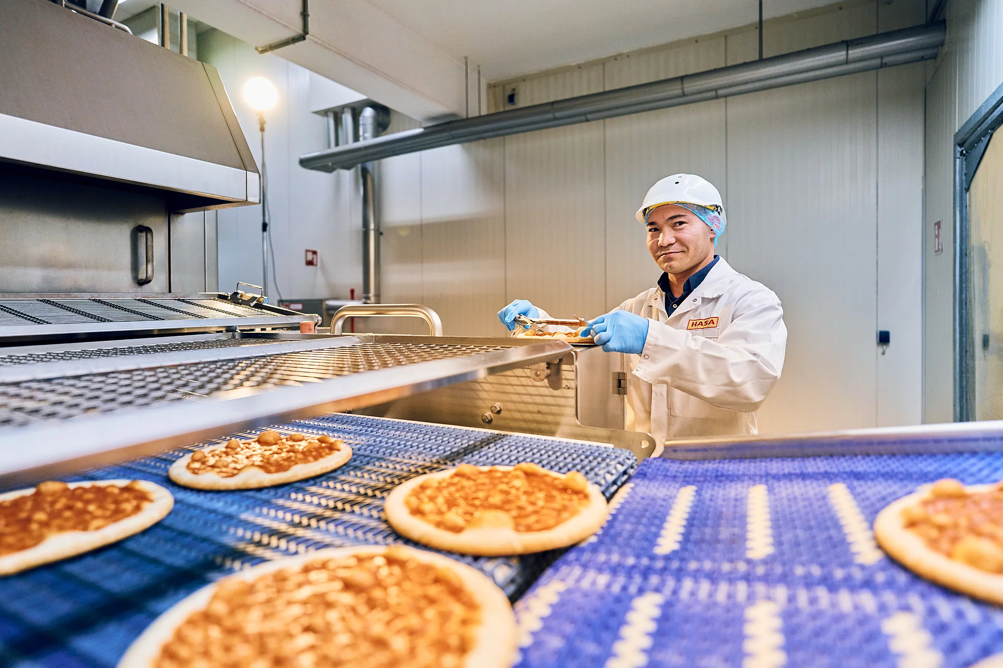 Mitarbeiterin in Arbeitskleidung vor einem Fließband mit Pizzen in der Produktion.