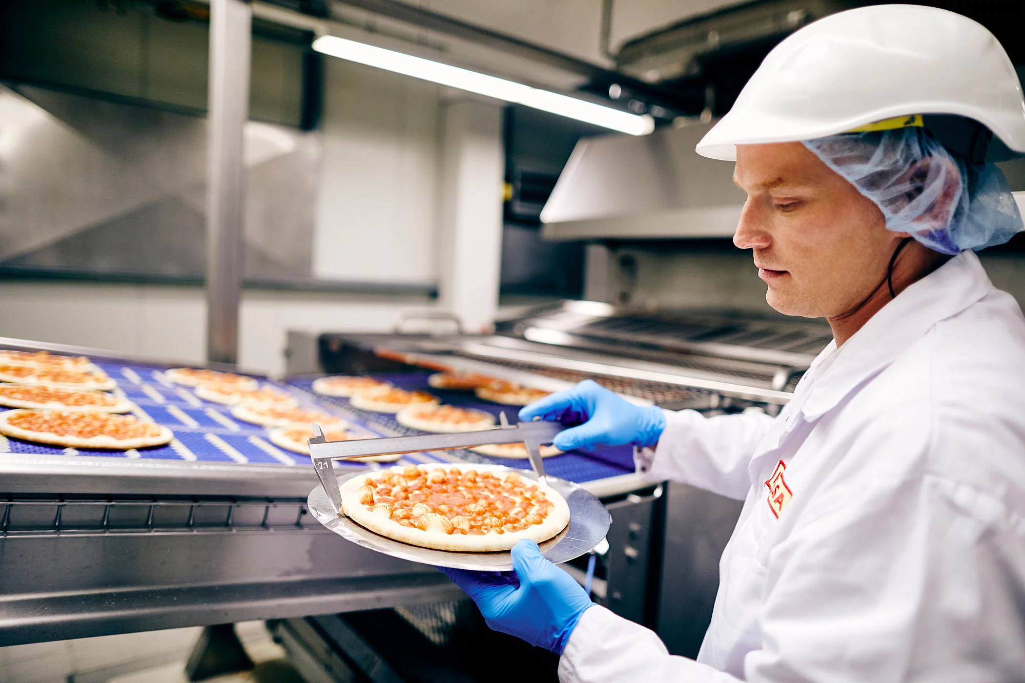 Mitarbeiter in Arbeitskleidung vor einem Fließband hält eine Pizza in der Hand.