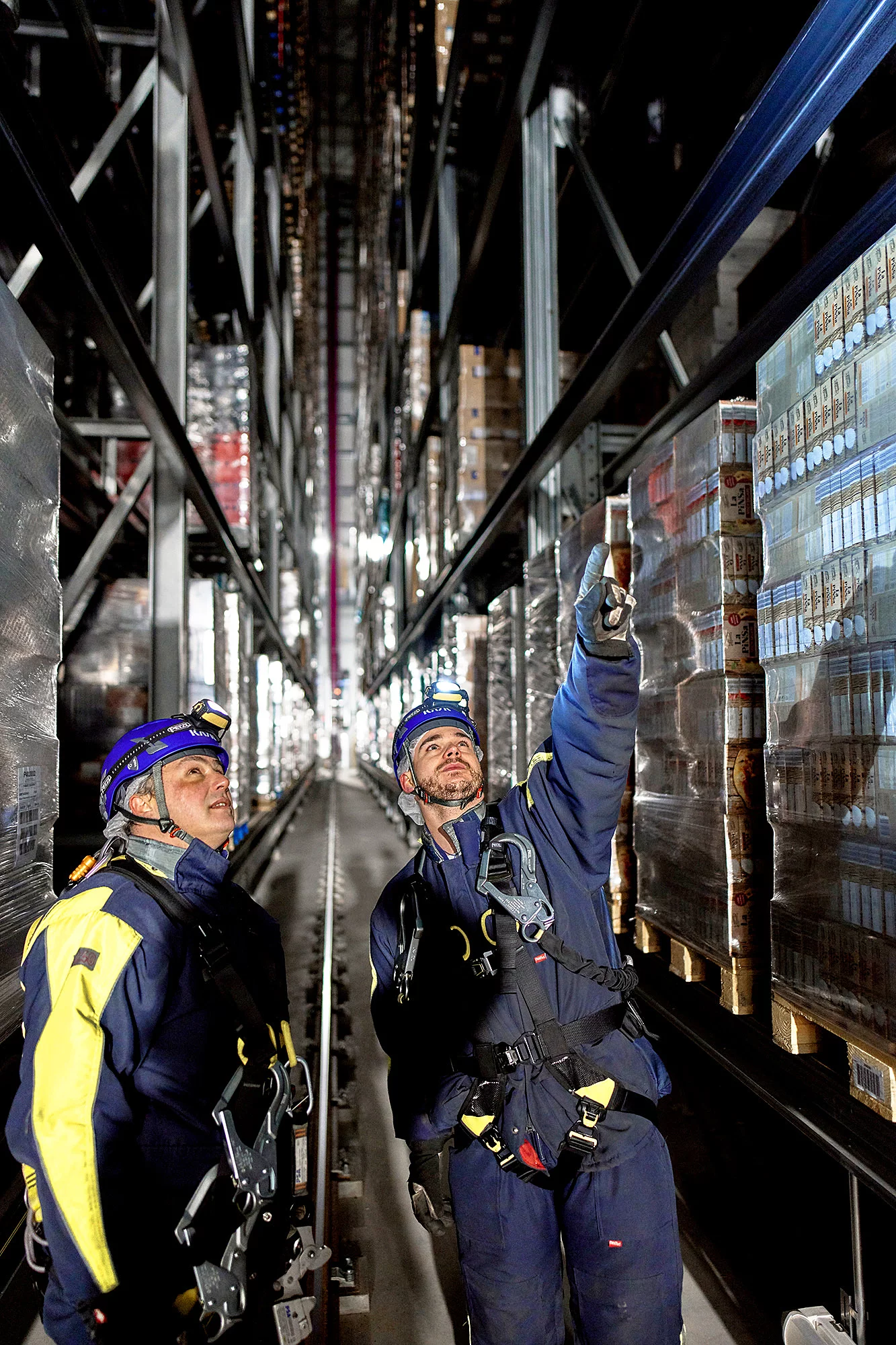 Zwei Mitarbeiter stehen in Arbeitskleidung im Hochregallager, einer zeigt nach oben.