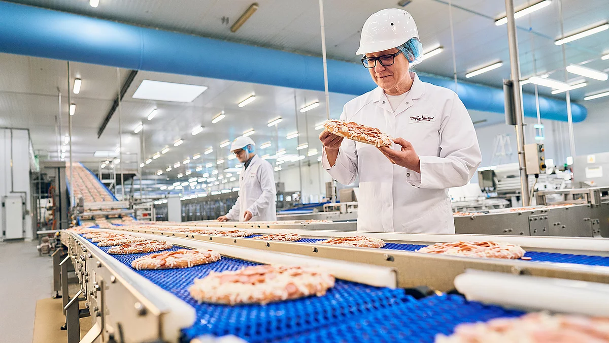 Mitarbeiterin in Arbeitskleidung steht am Pizza Produktionsband und hält eine Pizza in der Hand.