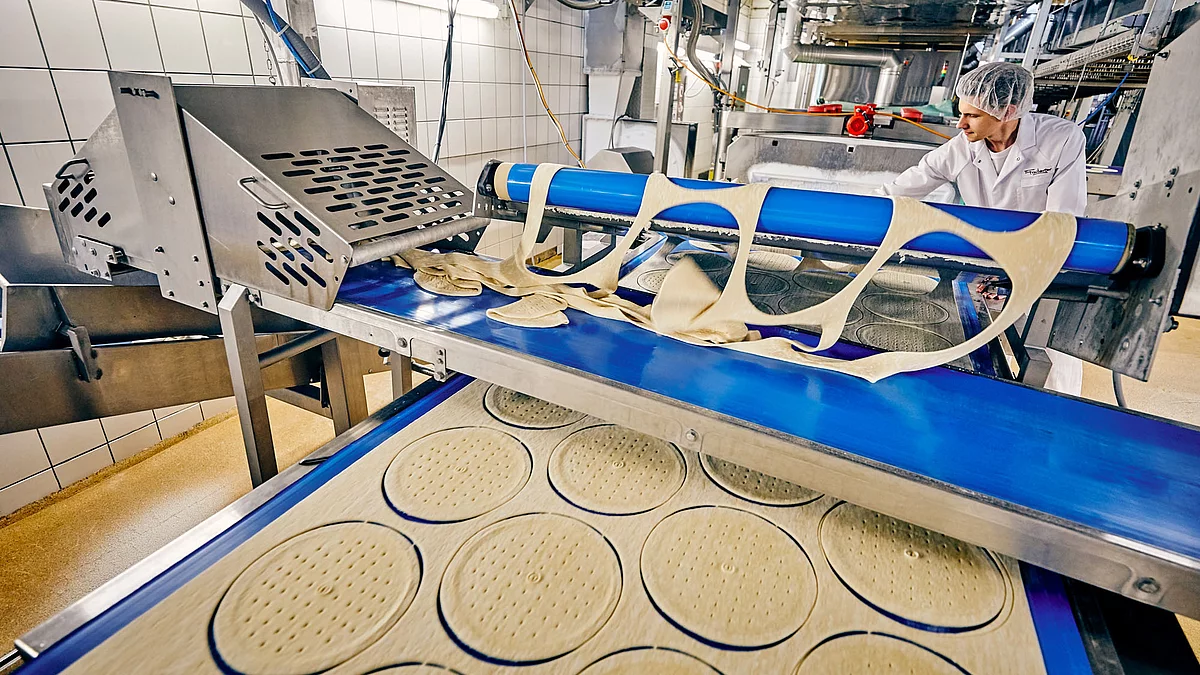 Mitarbeiter in Arbeitskleidung an Pizza-Produktionsmaschine.