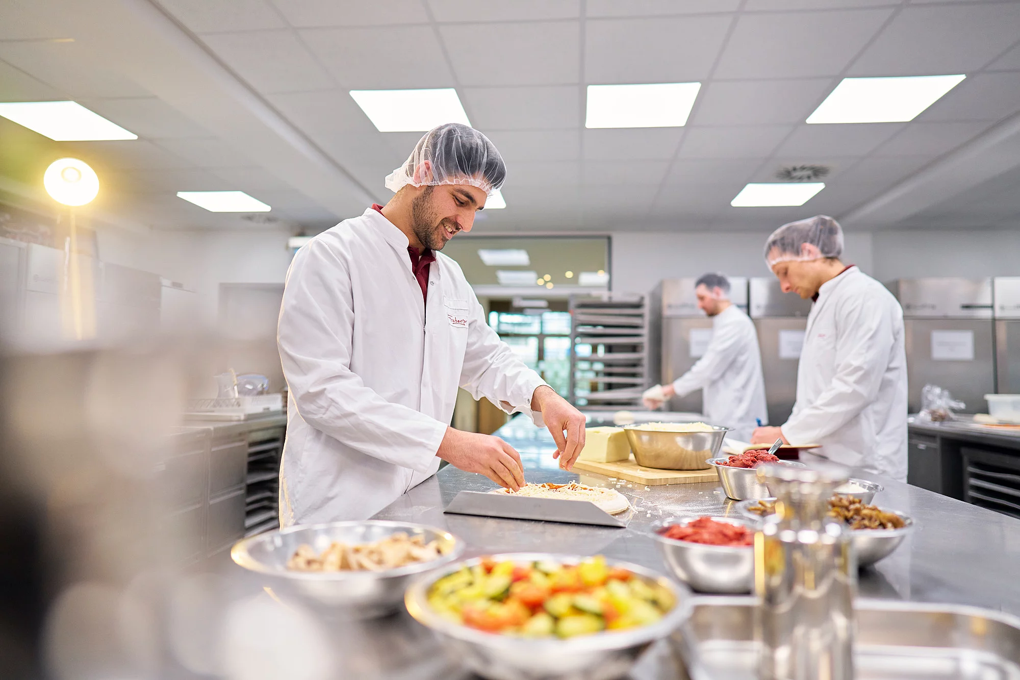 Drei Mitarbeitende in Arbeitskleidung in der Produktionsküche am Tresen kochen.