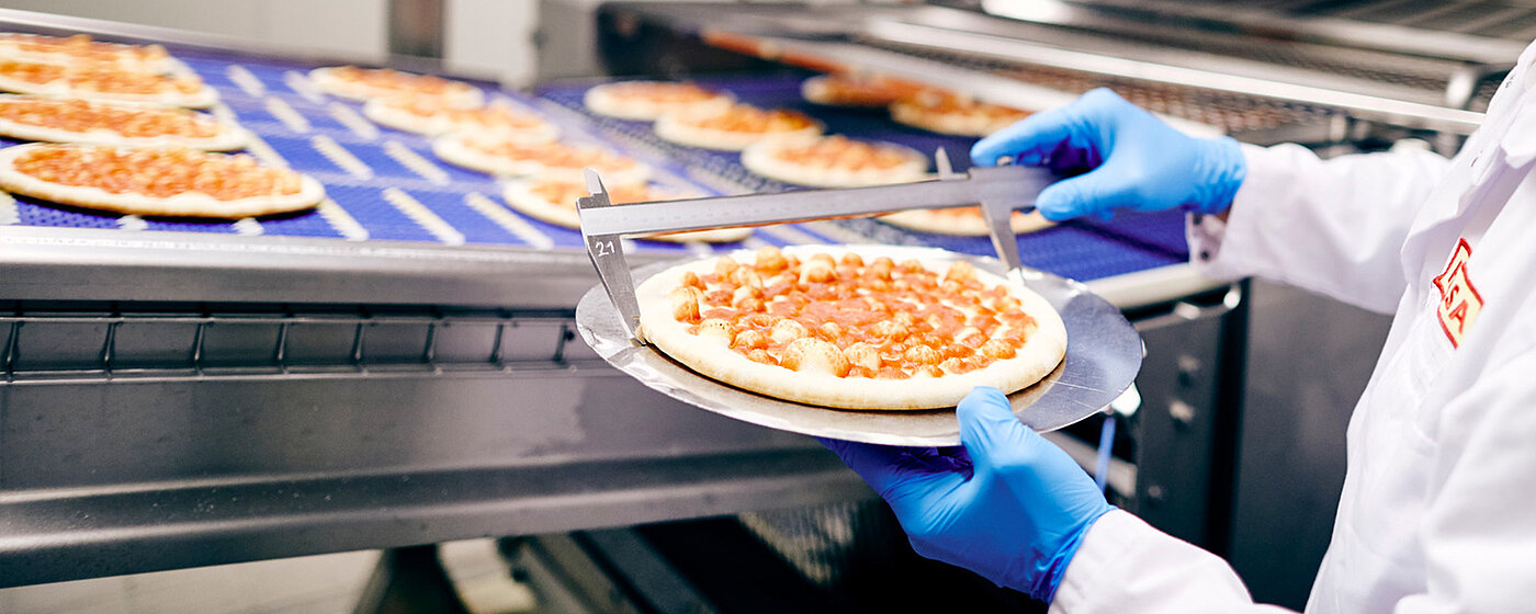 Pizzabefülltes Band. Mitarbeitende nimmt eine Pizza vom Band und misst diese.