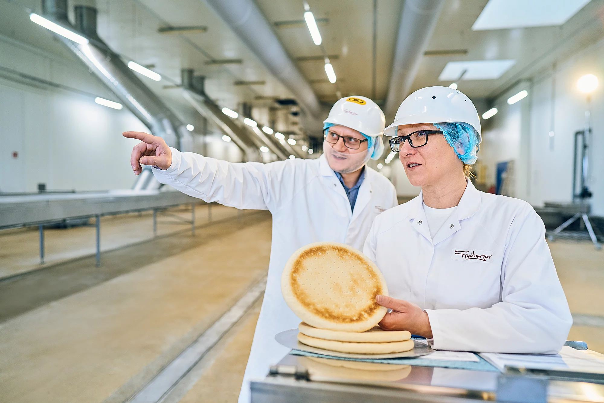 Zwei Mitarbeitende stehen in der Produktion. Die eine hält gebackenen Pizzateig in der Hand, der andere zeigt in eine Richtung.