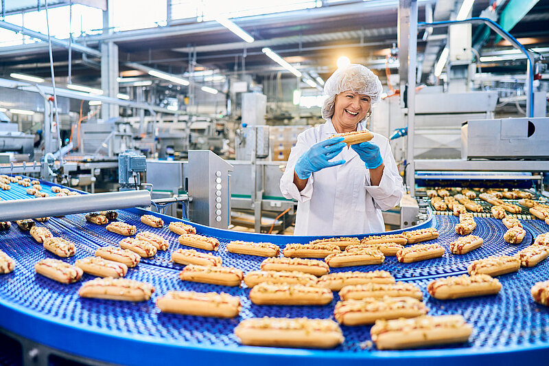 Lächelnde Mitarbeiterin mit Hotdog in der Hand. Vor ihr ein Fließband mit Hotdogs.