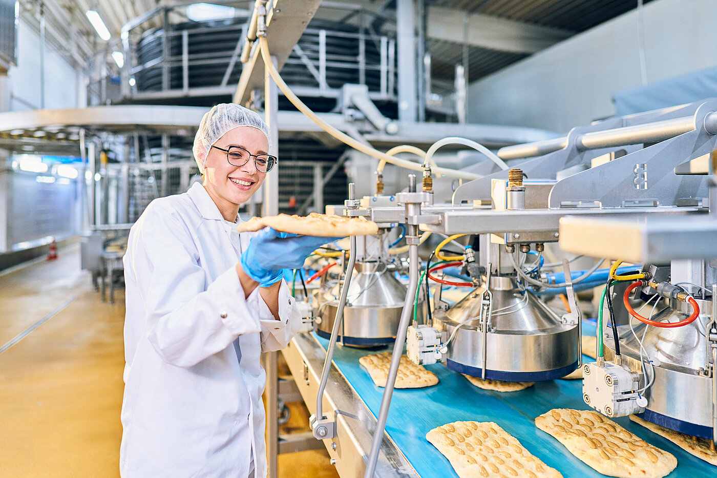 Mitarbeiter in Arbeitskleidung mit Pizza-Baguette in der Hand an der Produktionsmaschine.