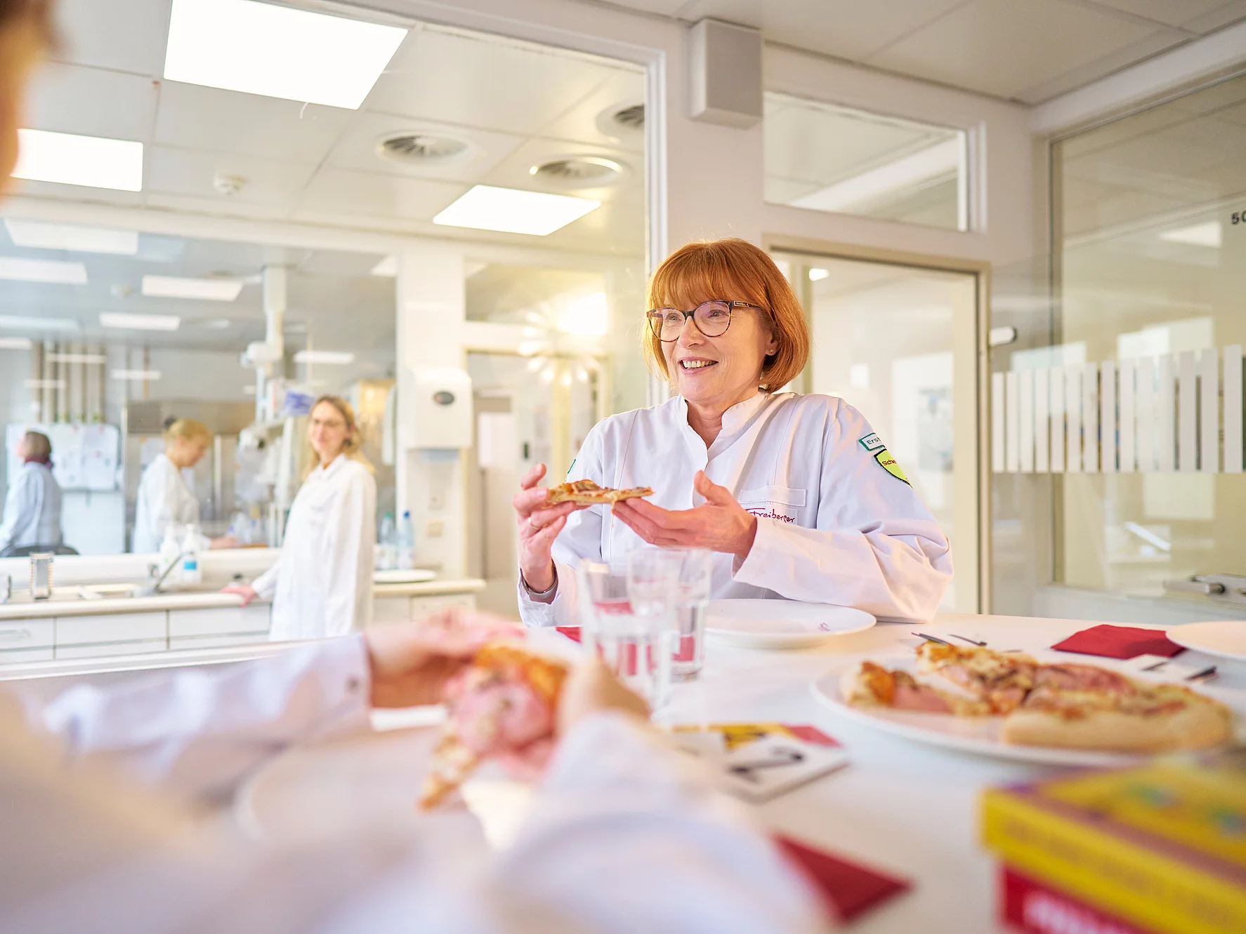 Zwei Mitarbeitende stehen am Tisch und halten ein Stück Pizza in der Hand. Im Hintergrund sieht man weitere Mitarbeitende.