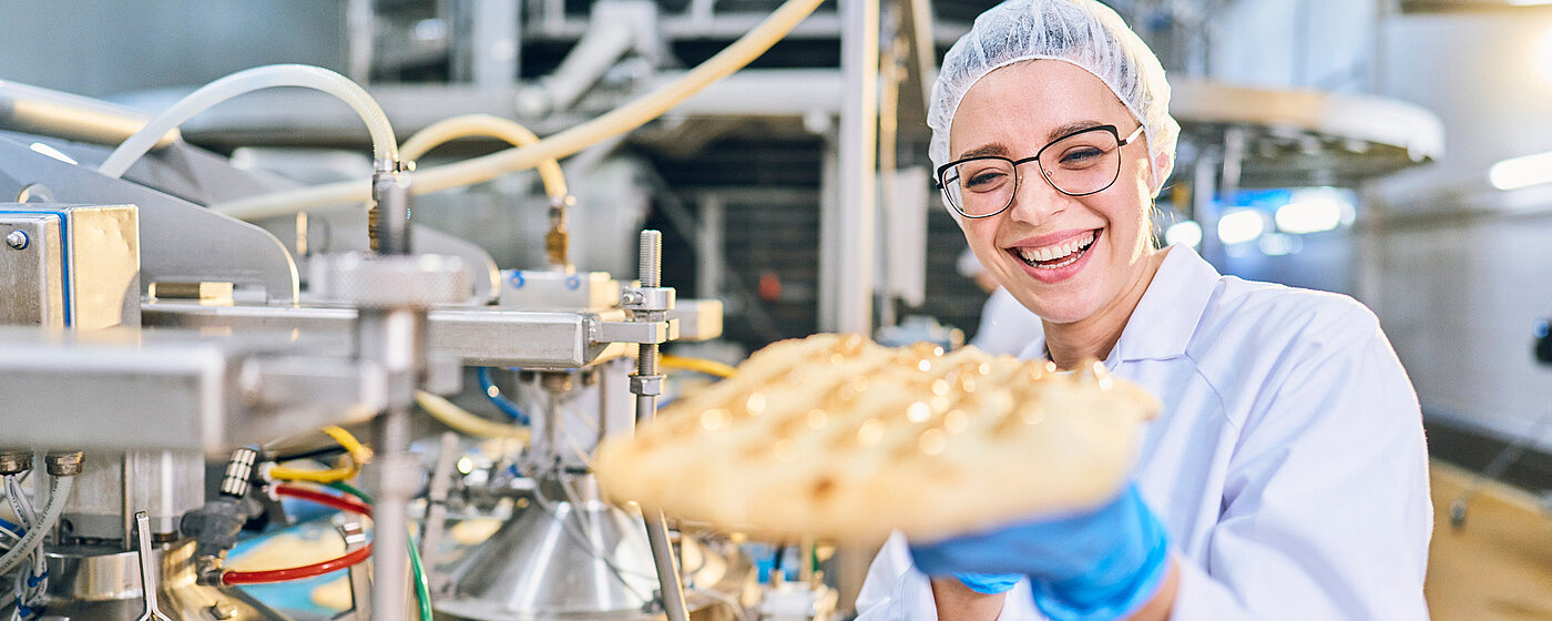 Eine Mitarbeiterin in Arbeitskleidung hält lächelnd einen Pizzateig in der Hand.