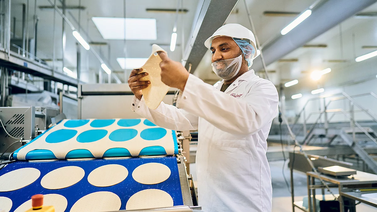 Mitarbeiter in Arbeitskleidung hält am Pizza-Produktionsband einen Pizzaboden in der Hand.