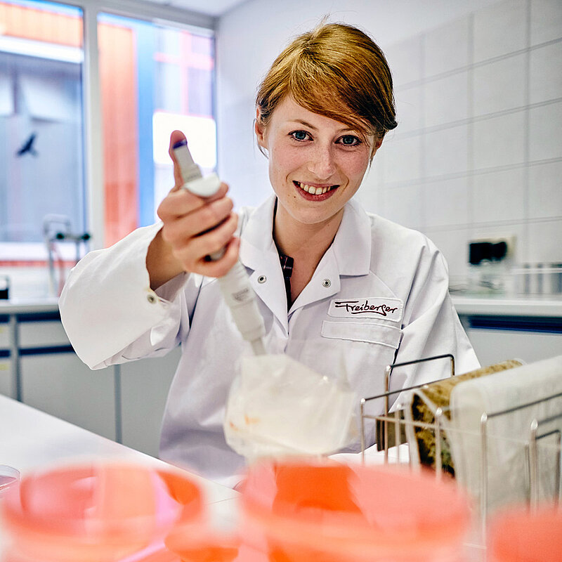 Eine Mitarbeiterin schaut lächelnd in die Kamera und hält eine Spritze in der Hand. 