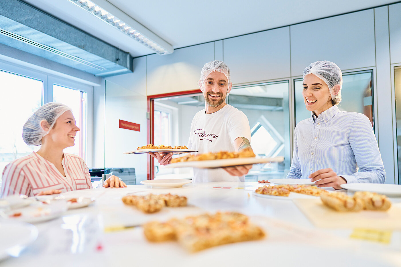 Drei Mitarbeitende stehen um einen Tisch voller Pizza, einer hält zwei Pizzableche in der Hand.