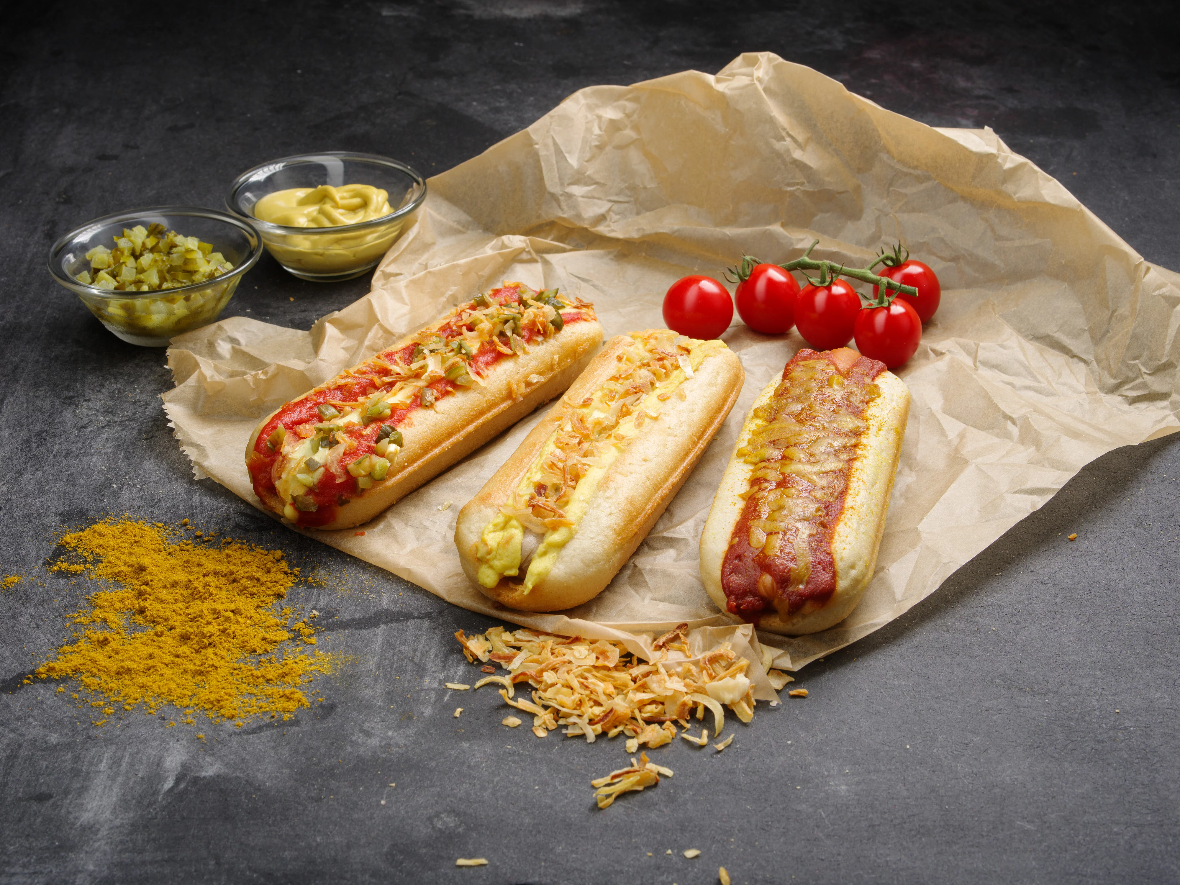 Drei Hotdogs auf einem Backpapier mit Röstzwiebeln, Tomaten und Gurken dekoriert.