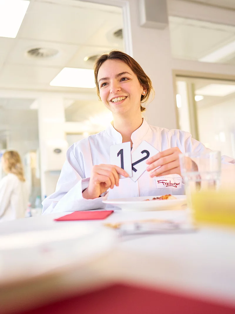 Eine Mitarbeiterin steht vor einem Tisch und hält zwei Kärtchen mit einer 1 und einer 2 darauf in der Hand. Im Hintergrund befinden sich weitere Mitarbeitende.