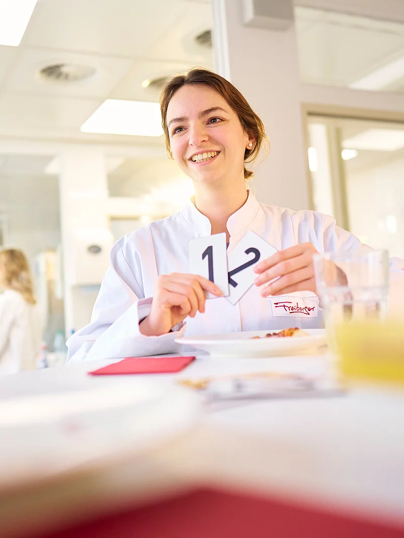 Eine Mitarbeiterin steht vor einem Tisch und hält zwei Kärtchen mit einer 1 und einer 2 darauf in der Hand. Im Hintergrund befinden sich weitere Mitarbeitende.