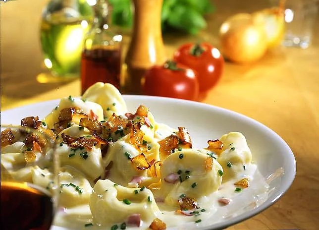 Tortelloni mit Sahnesoße auf einem Teller angerichtet, dahinter dekoriert mit Tomaten, Zwiebeln und Pfeffermühle.
