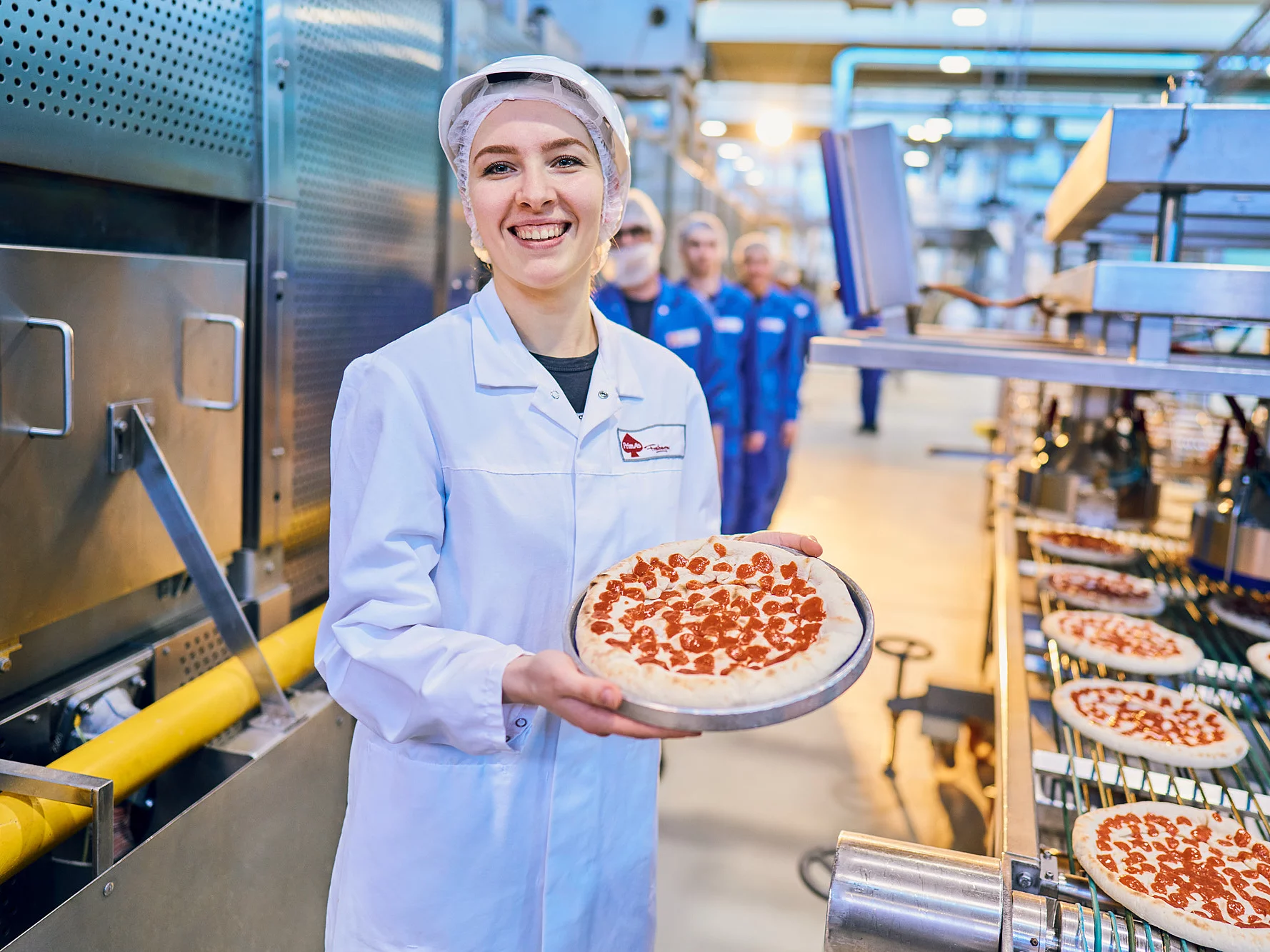Mitarbeiterin in Arbeitskleidung mit Pizza in der Hand an dem Produktionsband.