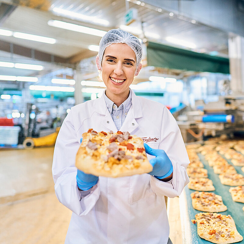 Eine Mitarbeiterin hält lächelnd ein Pizzabrot in der Hand.