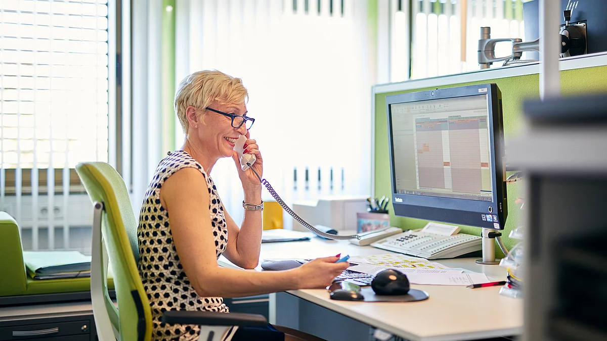 Mitarbeiterin im Büro am Schreibtisch mit einem Telefon in der Hand. 