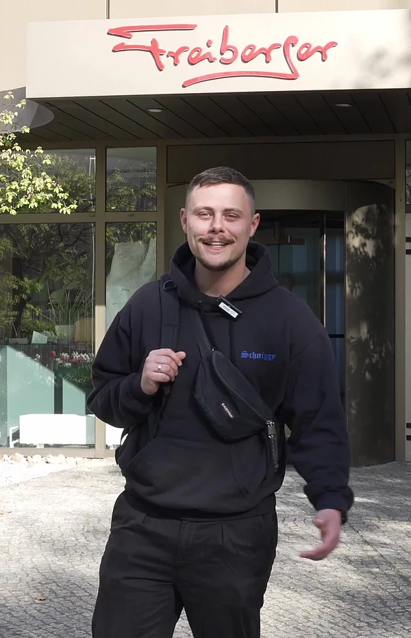 Person steht mit Mikrofon in der Hand draußen vor einem Eingang mit Freiberger Logo.
