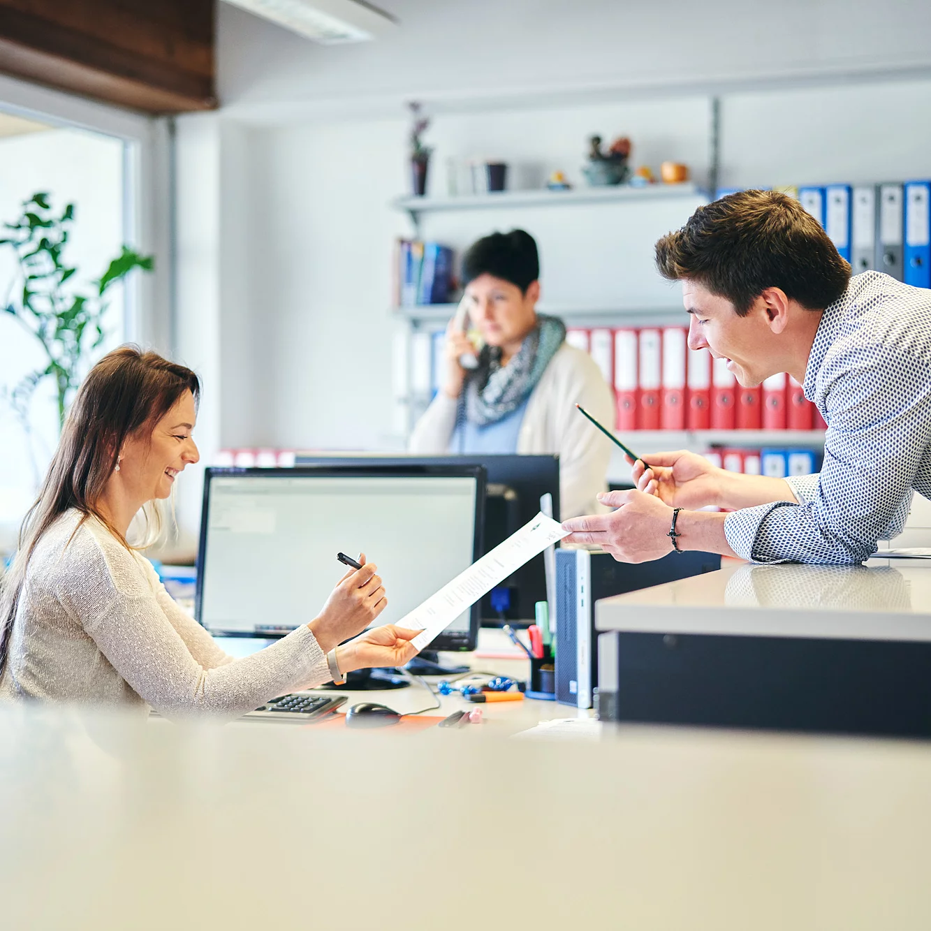 Drei Mitarbeitende stehen in einem Büro, zwei lächeln sich an und reichen sich Papier und Stift.