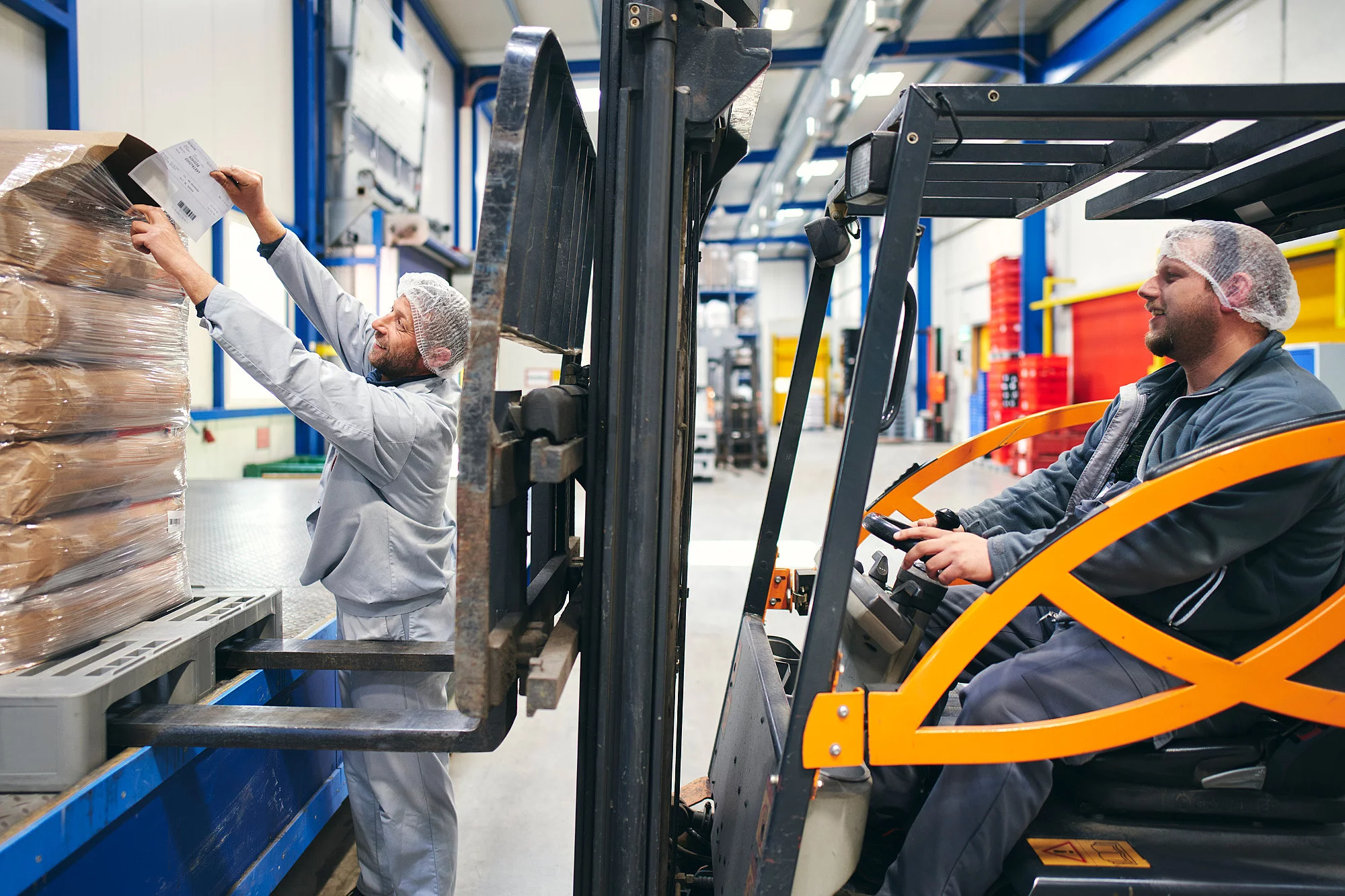 Zwei Mitarbeiter in der Produktion. Einer sitzend auf einem Gabelstapler, der andere stehend neben der Ware mit einem Zettel in der Hand.