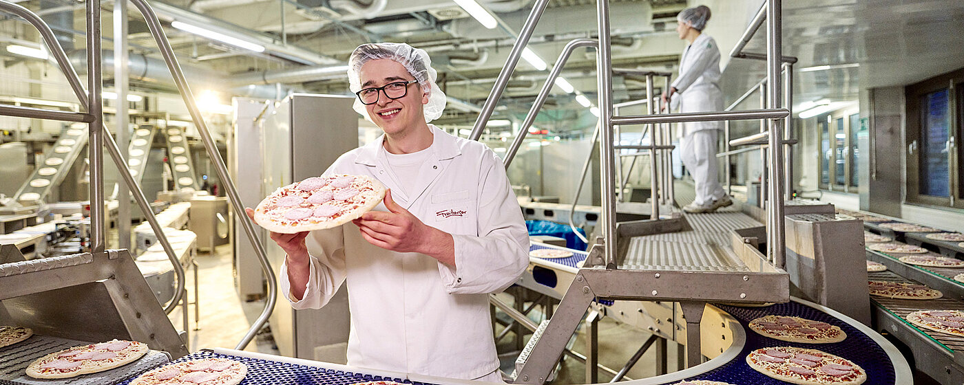 Mitarbeitender vor der Produktionsmaschine hält eine Pizza, vom Produktionsband, in den Händen.