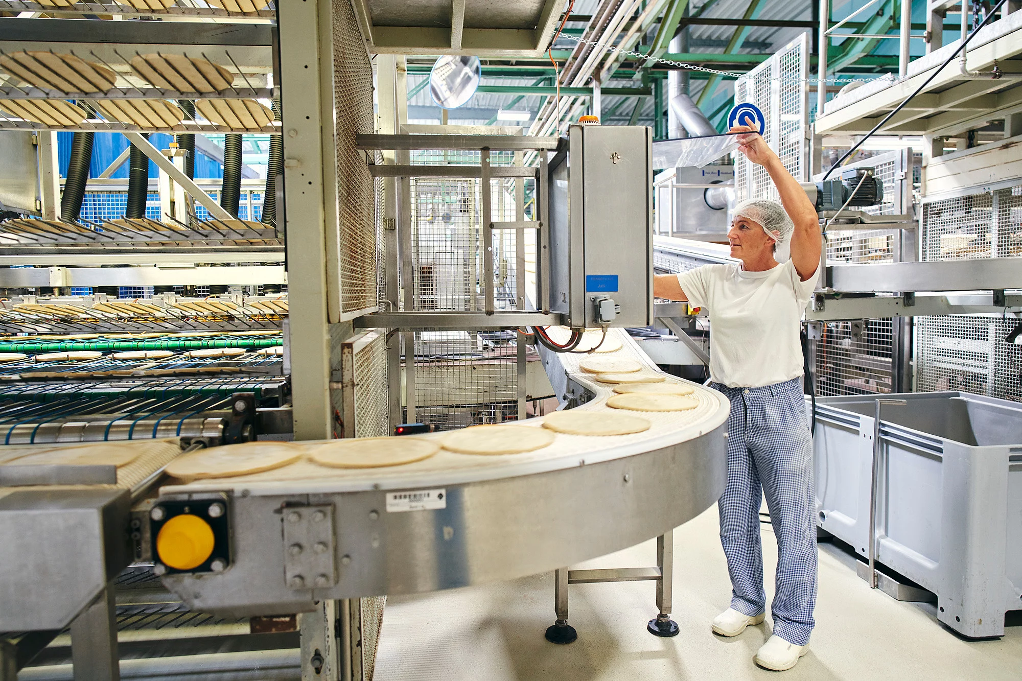 Mitarbeiterin in der Produktion an einem Fließband, befüllt mit Pizzateigen.